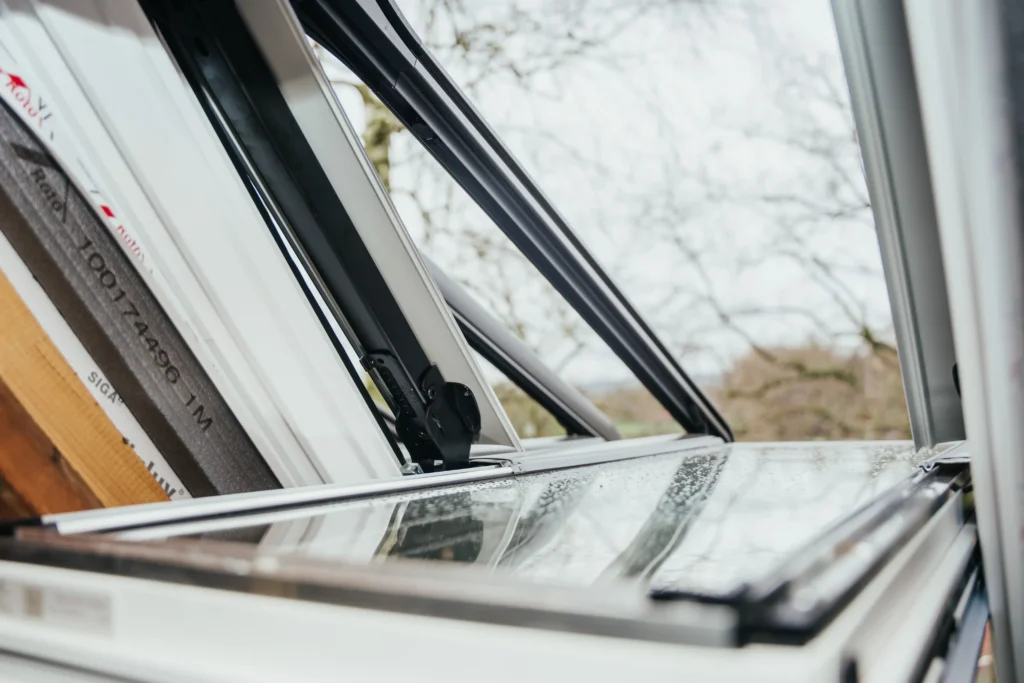 Geöffnetes Dachfenster mit metallischen Rahmen und Wassertropfen auf Glas, unscharfe Bäume im Hintergrund.