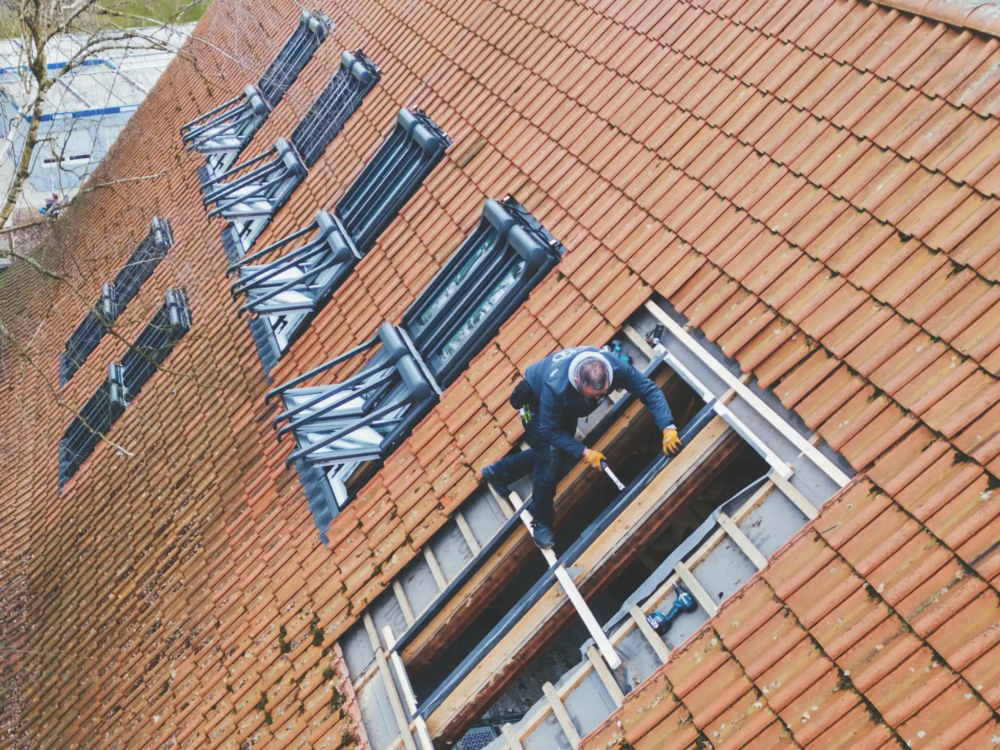 Ein Arbeiter repariert ein Dachfenster auf einem geneigten Ziegeldach in ruhiger, konzentrierter Atmosphäre.