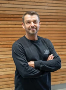 Person in schwarzem Oberteil mit Logo steht selbstbewusst vor Holzwand, Arme verschränkt, Blick direkt.