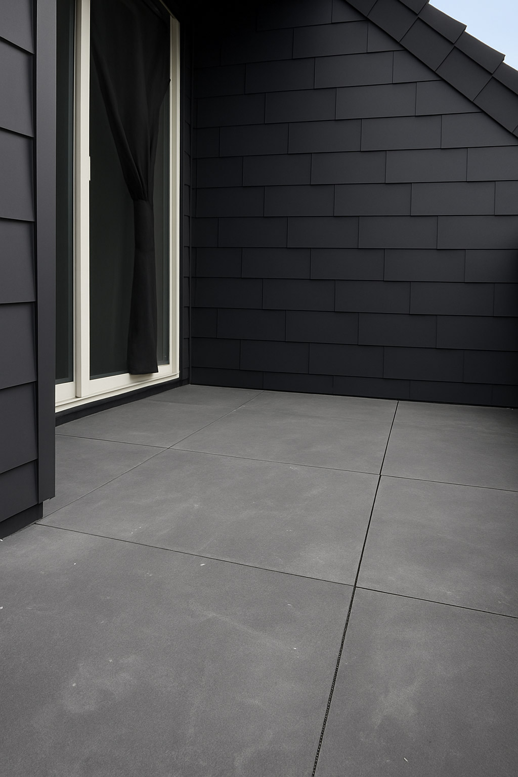 Moderne Terrasse mit grauen Bodenplatten, dunkler Schindelwand und Glastür mit schwarzem Vorhang.