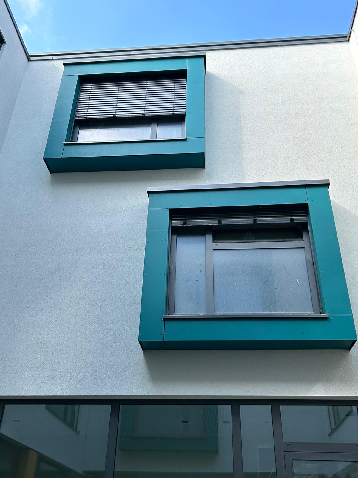 Modernes Gebäude mit zwei türkis umrahmten Fenstern; obere Jalousien geschlossen, untere offen.