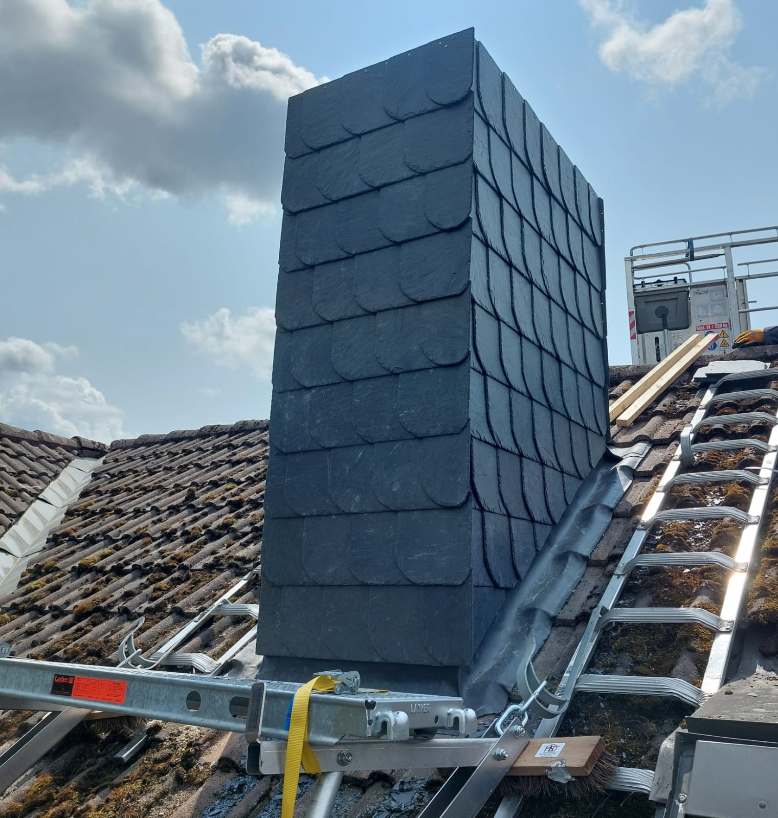 Ein Dach mit moosigen Ziegeln, ein großer Schornstein aus Schiefer und ein Baugerüst bei klarem Himmel.